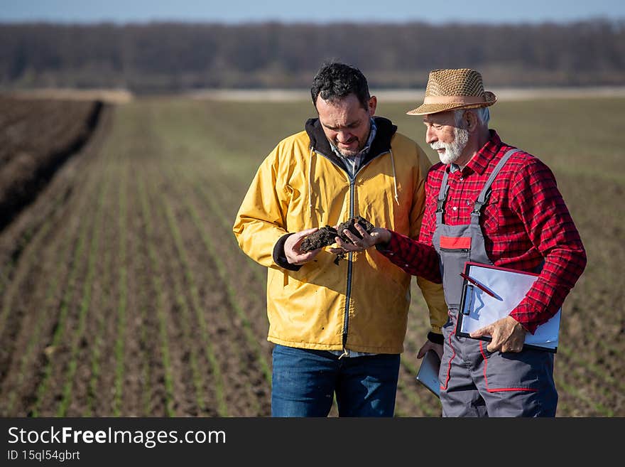 Two farmers checking soil quality in field, senior man holding notepad, while younger carrying lump of earth
