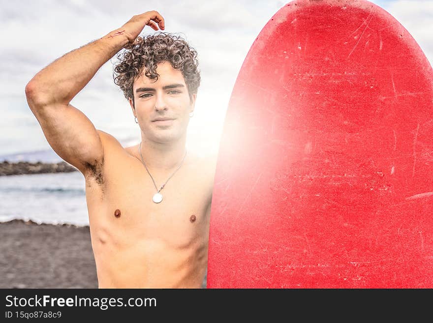 Young handsome spanish man with curly hair standing shirtless on the beach with red surfboard, smiling to the camera. Good vibes