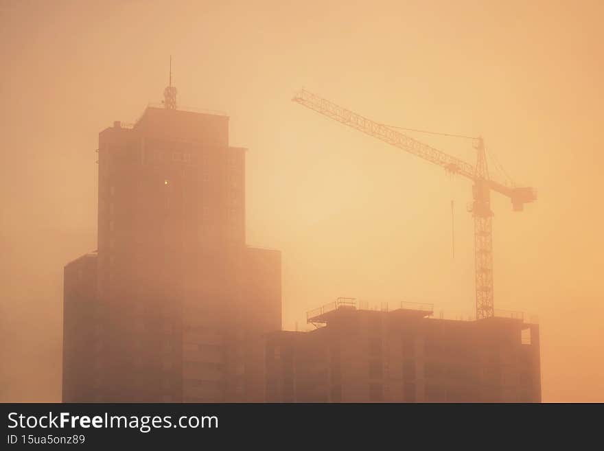 Silhouette of a construction crane and unfinished residential building on a foggy autumn morning during the sunrise. Housing