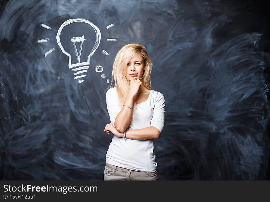 Beautiful young smart blonde woman is thinking near the chalk board with a drawn light bulb. Creative idea concept