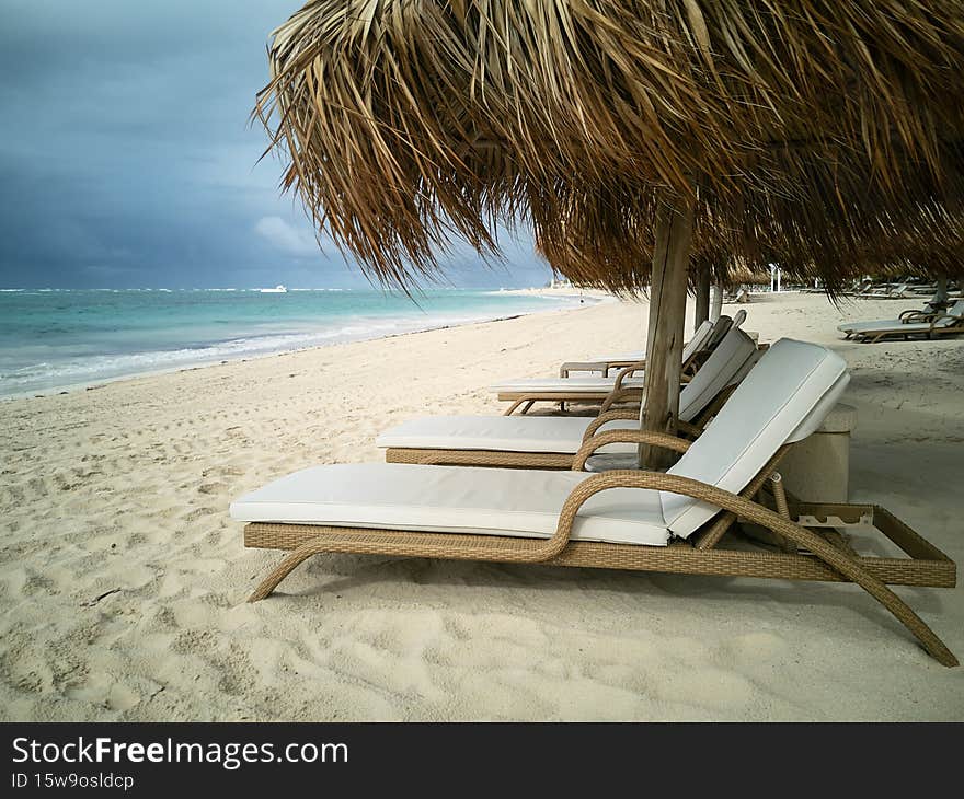 Sun loungers on ocean beach under umbrella from palm branches. Sandy beach with no people