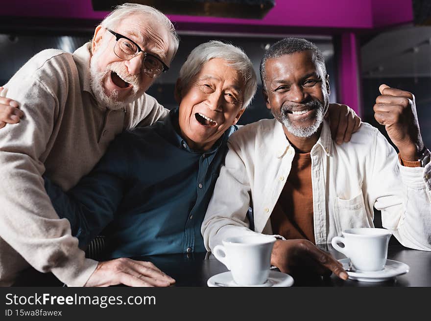 smiling african american man showing rejoice gesture near laughing senior friends in cafe,stock image