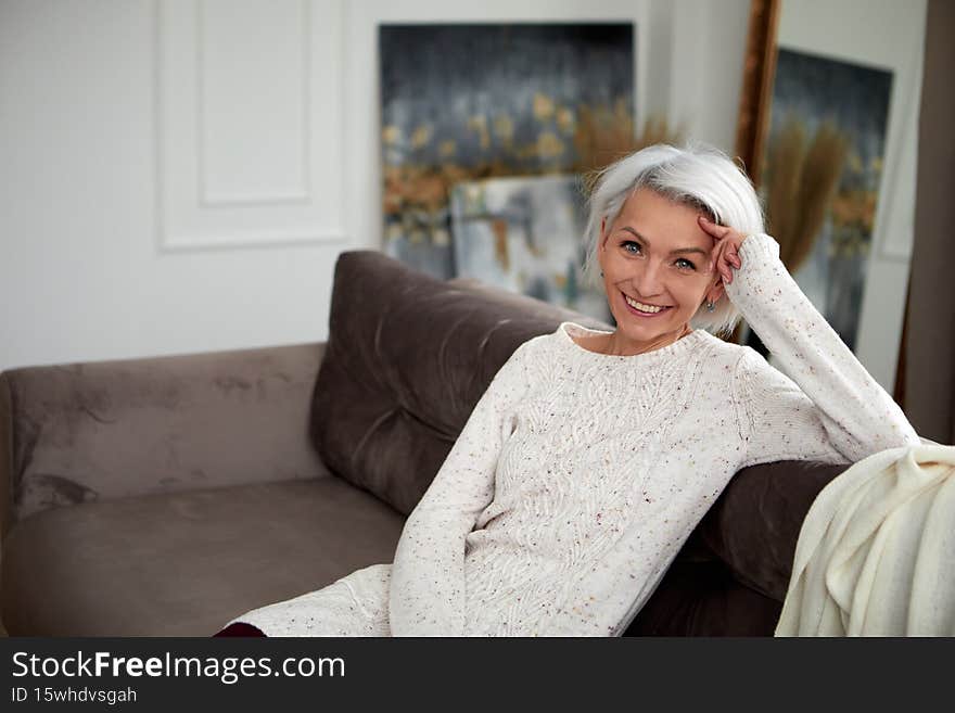 Charming mature female in knitted sweater and with gray hair sitting on comfortable sofa and leaning on hand while looking at camera with smile. Charming mature female in knitted sweater and with gray hair sitting on comfortable sofa and leaning on hand while looking at camera with smile