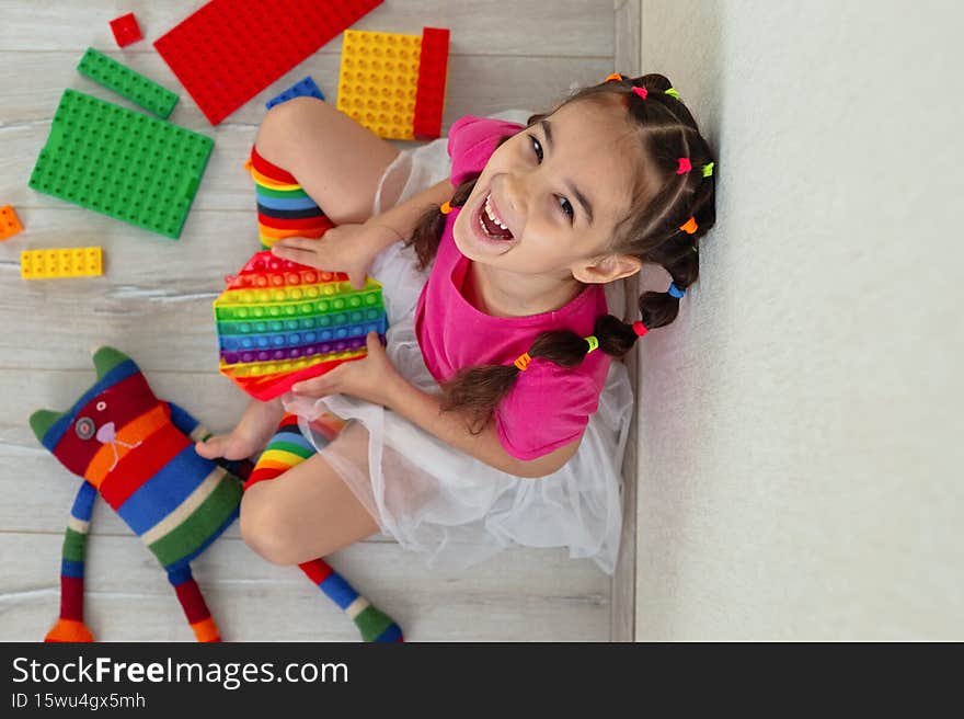 cheerful little brunette girl in bright colorful clothes, sits at home, on the floor