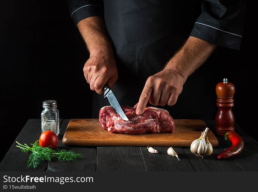 Cook cuts meat with a knife in the kitchen prepares food. Vegetables and spices on the kitchen table in a restaurant to prepare a