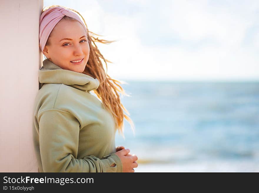 Portrait of a young smiling woman with dreadlocks posing against a white wall. In the background is the ocean and the