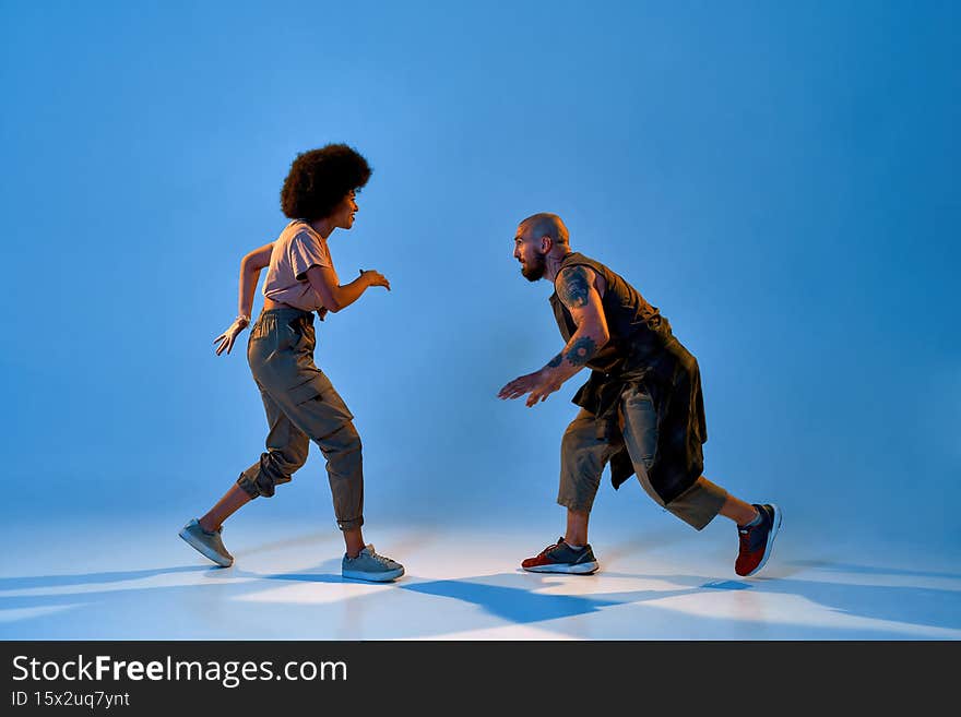 Mixed-race dance couple dancing breakdance. Concept of contemporary dancing. Idea of choreography. Flexible caucasian man and black girl on blue background in studio with shadow on floor. Copy space. Mixed-race dance couple dancing breakdance. Concept of contemporary dancing. Idea of choreography. Flexible caucasian man and black girl on blue background in studio with shadow on floor. Copy space