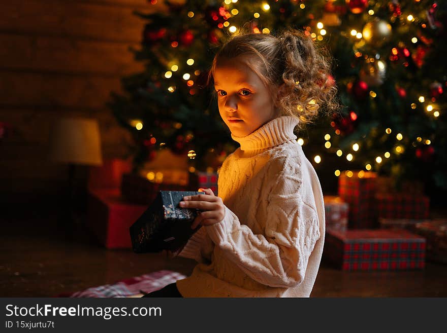 Pretty happy little blonde child girl opening Christmas box with gift sitting on floor in xmas morning in dark living room with festive interior, on background of decorated tree with bokeh lights.