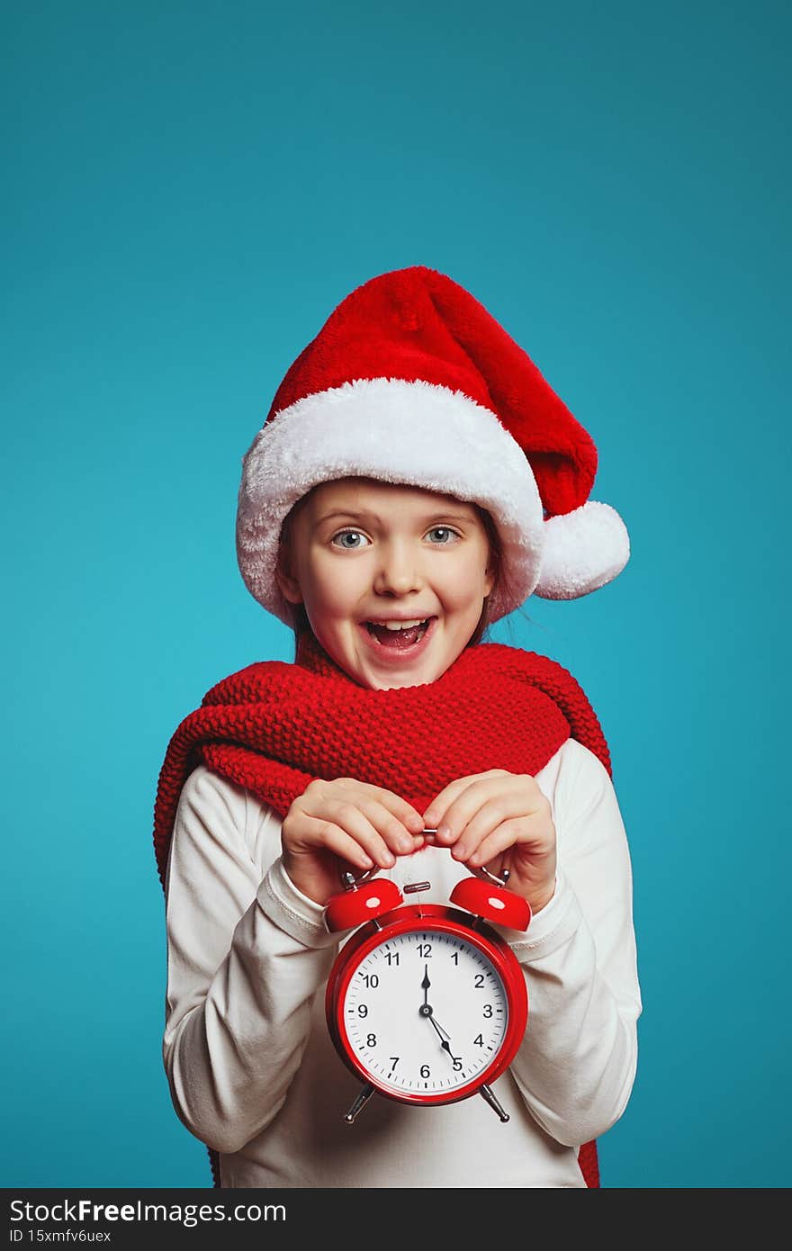 Vertical photo of amazed little caucasian kid, wearing christmas hat and red scarf holding alarm clock. Christmas night. Happy xmas and New Year. Vertical photo of amazed little caucasian kid, wearing christmas hat and red scarf holding alarm clock. Christmas night. Happy xmas and New Year