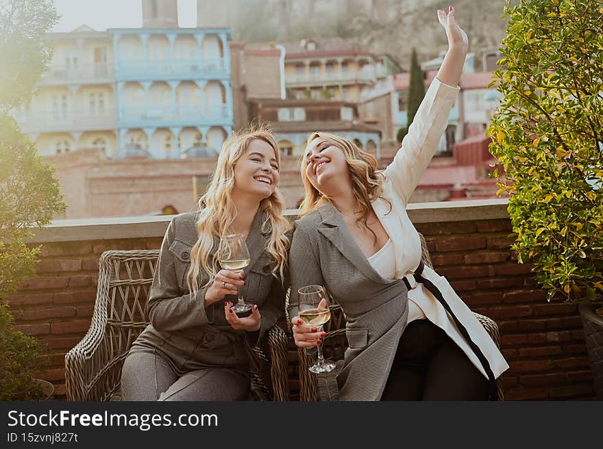 Close-up portrait of two female friends in strict suits laughing drinking wine on the terrace outside at summer street cafe on background buildings of Old Tbilisi city, Georgia. High quality photo