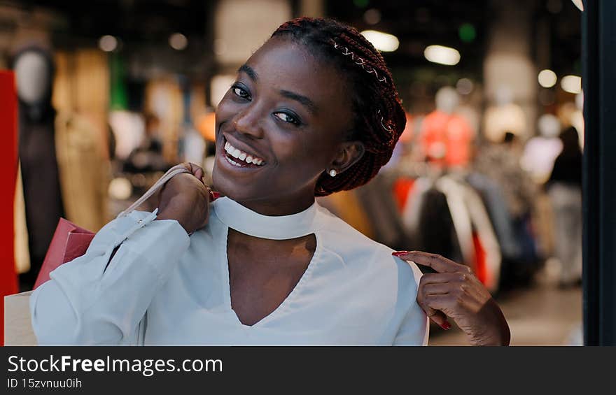 Smiling woman african american girl consumer buyer client stands indoors in store shopping mall shows at camera purchases happy with discount sale holds bags colored cardboard packages in hands. High quality photo