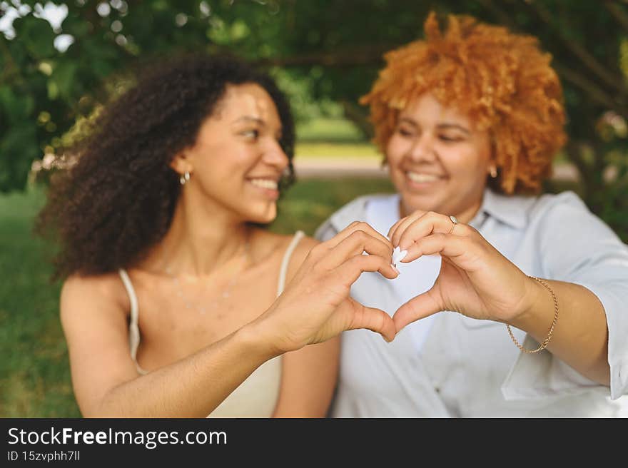 Young beautiful happy lesbian African American couple sitting on green grass showing love sign outside at nature summer