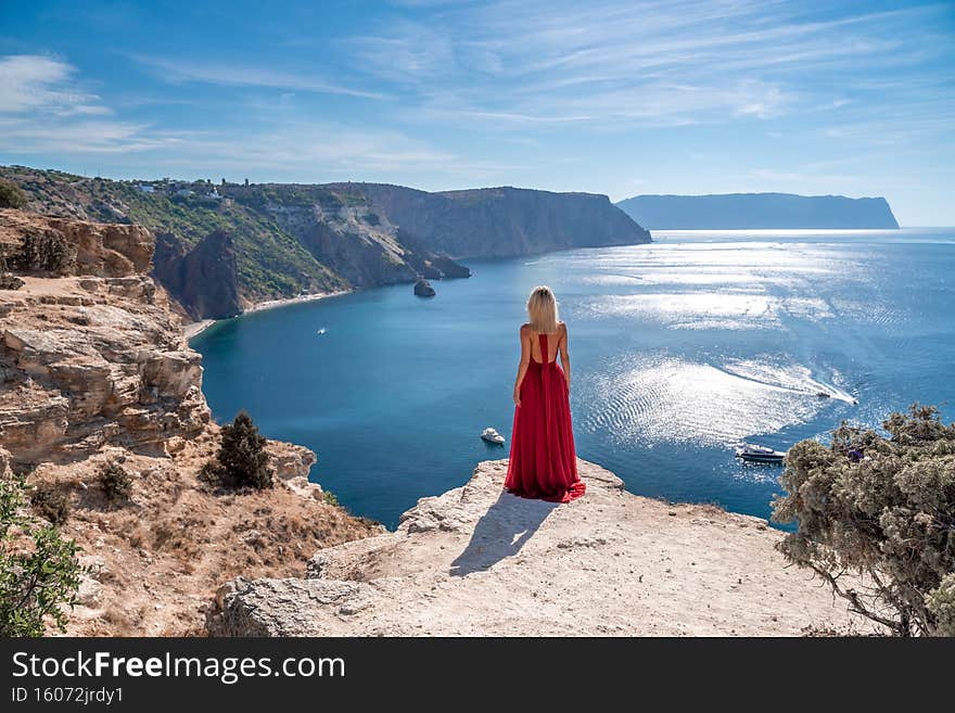 A girl with loose hair in a long red dress descends the stairs between the yellow rocks overlooking the sea. A rock can