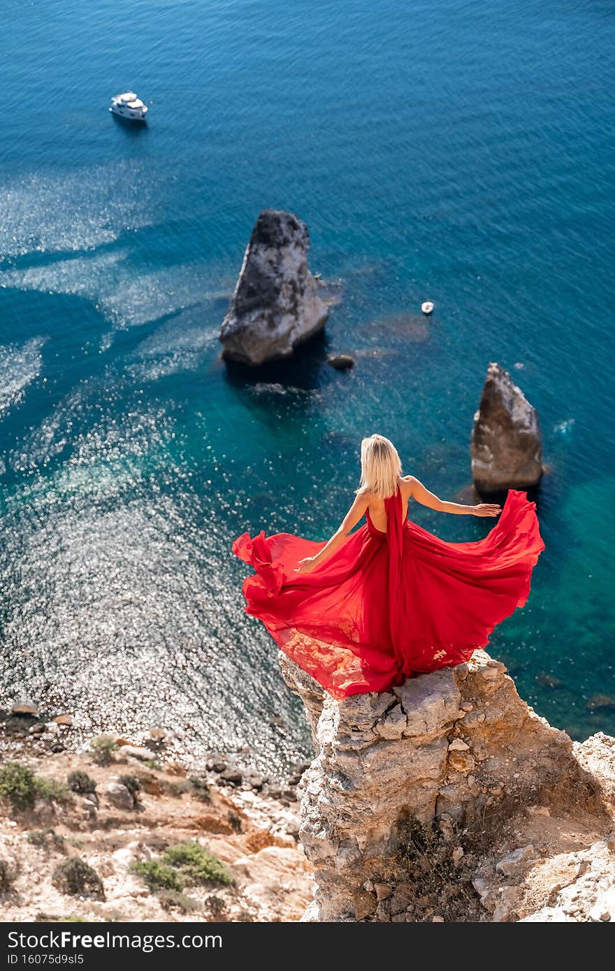 A girl with loose hair in a long red dress descends the stairs between the yellow rocks overlooking the sea. A rock can