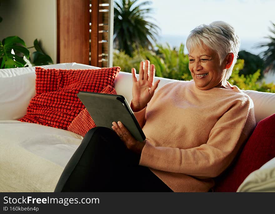 Multi-cultural elderly female waving at family on online call over her electronic tablet, sitting on living room sofa in daytime.
