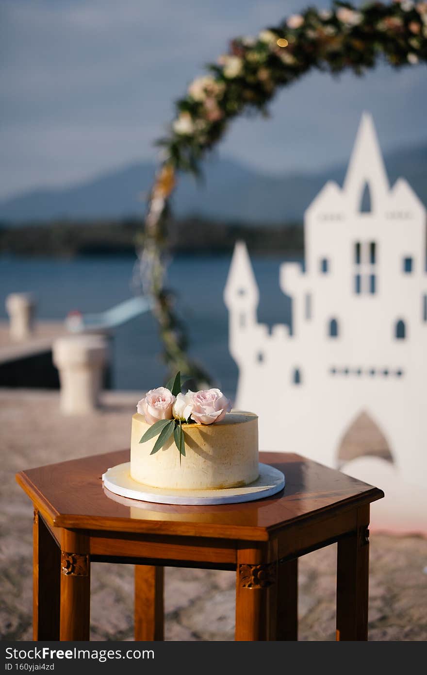 White cake with roses and green leaves, stands on a table against the background of a wedding arch