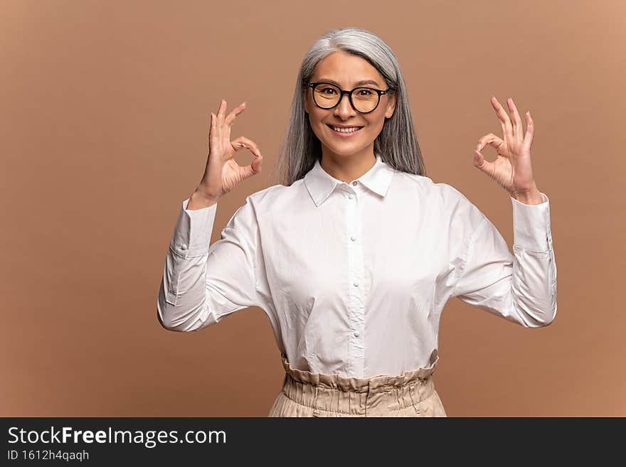 Portrait of satisfied asian senior woman with grey hair in formal white blouse standing, looking at camera showing Ok
