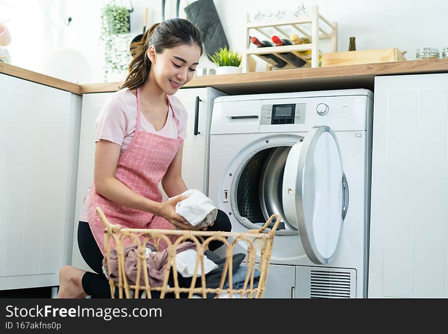 Asian beautiful woman put dirty clothes to washing machine in house. Attractive girl wear apron sit on floor, feel happy to loads laundry in washer appliance at home. Domestic-House keeping concept