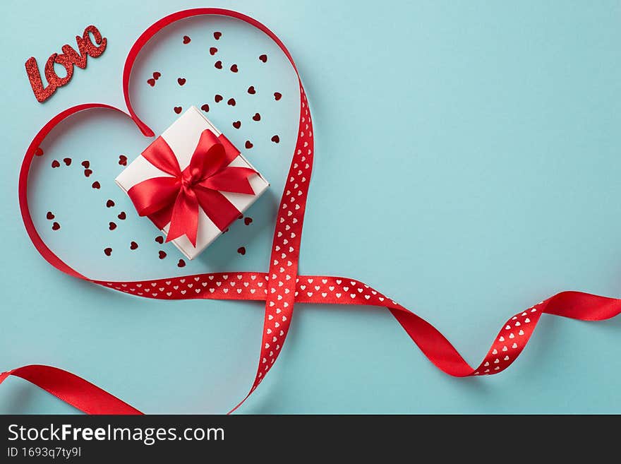 Top view photo of valentine`s day decorations white giftbox with red bow red silk ribbon heart inscription love and heart shaped confetti on isolated pastel blue background with blank space.