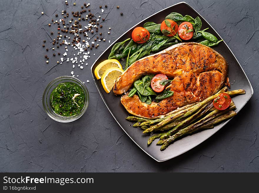grilled salmon steak, asparagus and fresh spinach with tomatoes on a black square plate on a dark background.