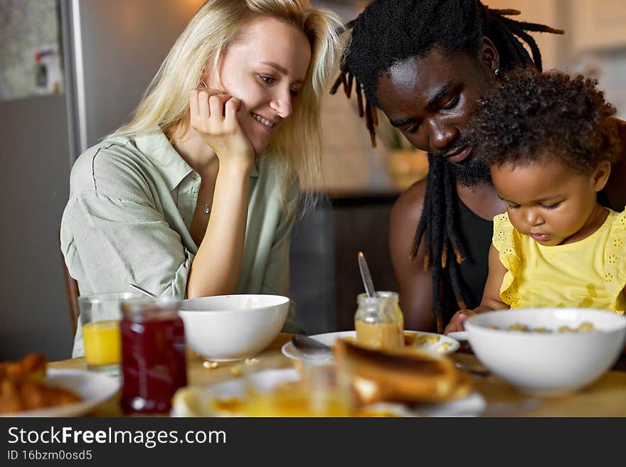 Diverse mother, father having breakfast with daughter sitting behind table with food, black child sit using smartphone watching cartoons, enjoying leisure, playing games on gadget. children concept