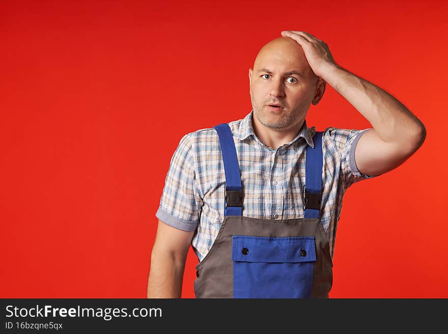 What have i done or fear and confusion, human emotions. Surprised and sad, a big bald man in overalls and a plaid shirt holds his head with his hand on a red background.