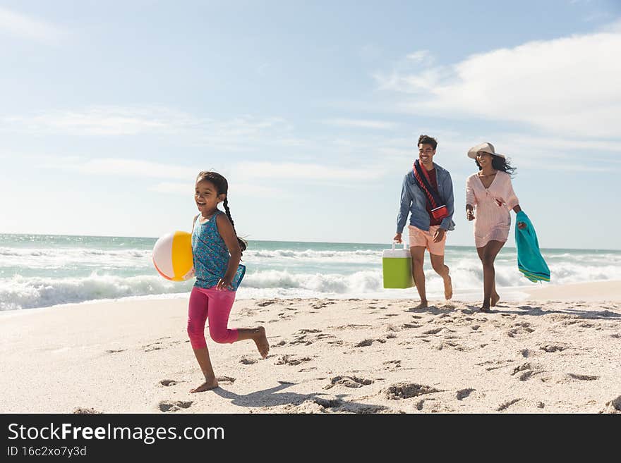 Full length of biracial girl with ball running ahead from parents on sand at beach against sky