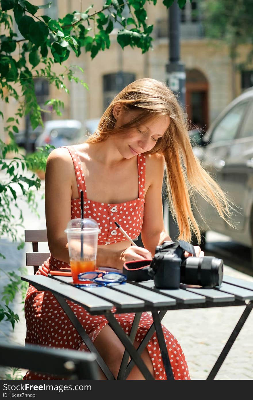 Happy young caucasian,millennial or gen z woman with long brunette holding pen in hand,making notes in modern outdoor cafe.Hipster Influencer using social networks,camera on table.