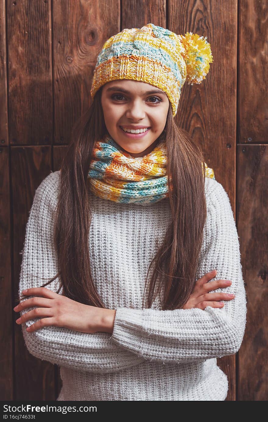 Portrait of a cute teenage girl in a scarf and a cap looking in camera and smiling, standing on a wooden background