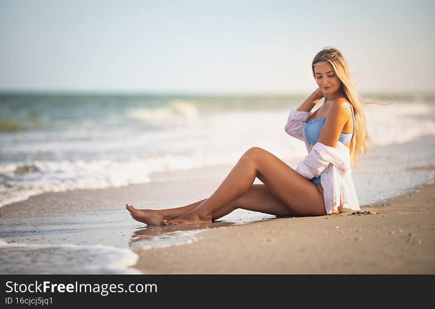 A girl in a bluish swimsuit and shirt sits near the water edge and peers into the horizon