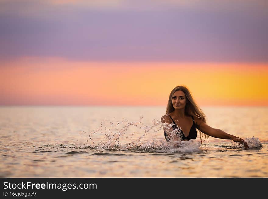 A slender tanned young girl with blond lush hair in a black swimsuit, splashes to the sides while sitting in a shallow estuary against the background of a bright warm sunset. A slender tanned young girl with blond lush hair in a black swimsuit, splashes to the sides while sitting in a shallow estuary against the background of a bright warm sunset