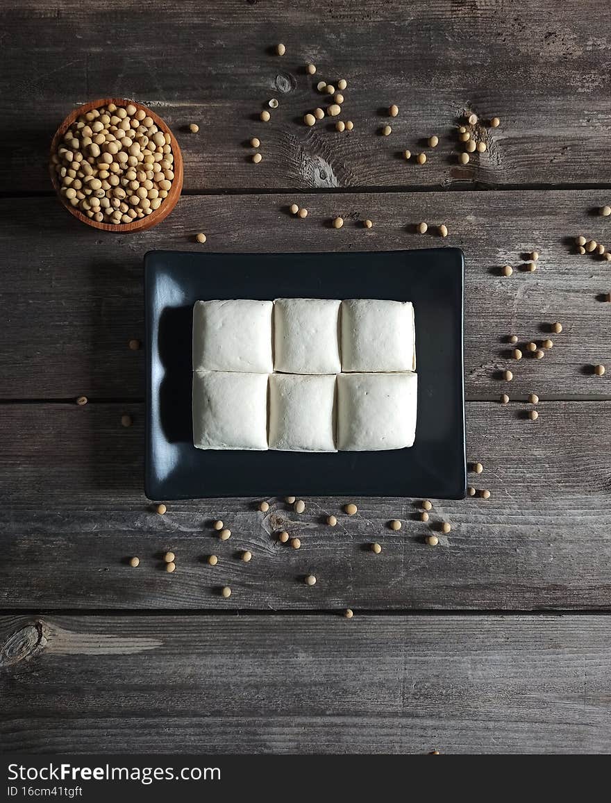 Tofu. Close view on fresh Tofu on black square plate, with rustic table background, a cup of raw soybean in the small wooden bowl and some grain mess on the base. Tofu is a food made from soy bean juice and native food from china. Healthy food for diet. Rich of Protein. Raw food. Tofu. Close view on fresh Tofu on black square plate, with rustic table background, a cup of raw soybean in the small wooden bowl and some grain mess on the base. Tofu is a food made from soy bean juice and native food from china. Healthy food for diet. Rich of Protein. Raw food.