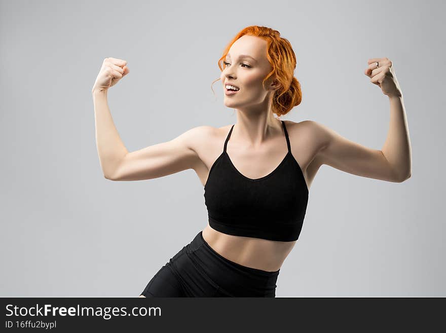 Self assured young fit female model with ginger hair in black sportswear smiling and showing biceps while standing against gray background and looking away. Self assured young fit female model with ginger hair in black sportswear smiling and showing biceps while standing against gray background and looking away