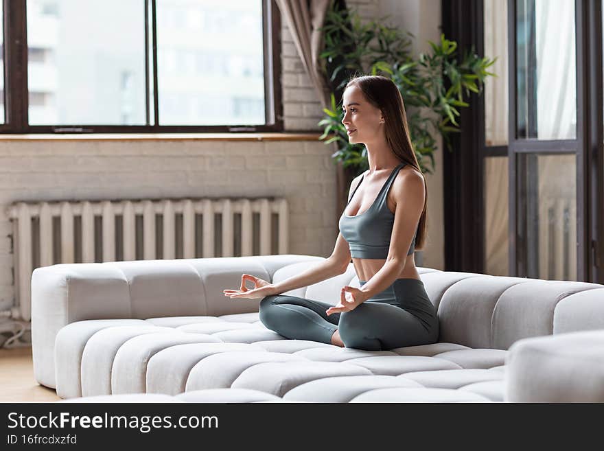 Beautiful healthy woman in a sports sportswear meditates on the couch at home, to be in harmony with body and soul