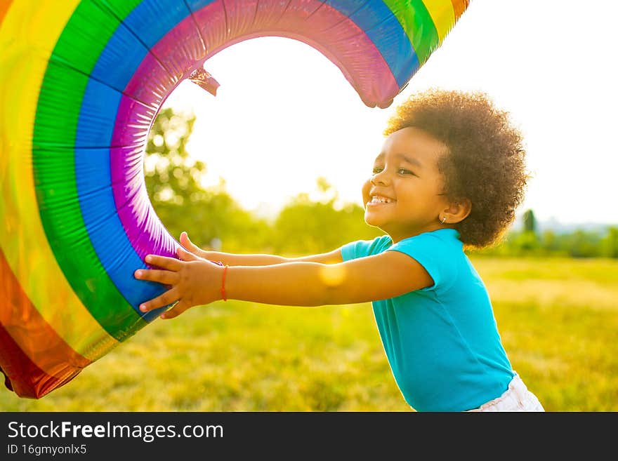 positive multicultural race little girl with afro curly hair holding rainbow balloon in summer park.