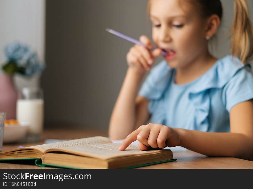 Close-up focus on foreground of thoughtful elementary child school girl doing homework and holding pen against mouth sitting at home table, selective focus. Homeschooling concept.