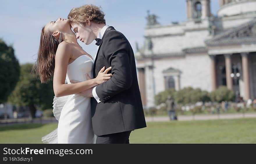 Beautiful newlyweds. Action. A couple of lovers, the bride in a tight white dress and the groom in a suit with long hair pose on the street next to the embankment and beautiful historical buildings in summer