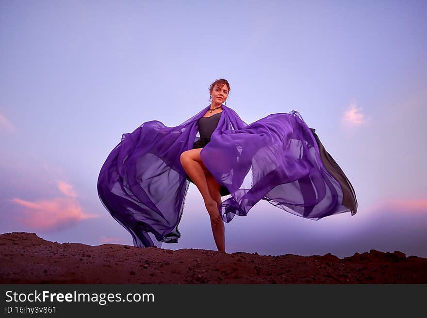 Beautiful girl with black dress and violet fluttering cloth dancing on sand dune. Model or dancer posing in nature landscape