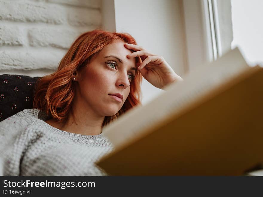 The girl with the orange hair is lying in bed. She is holding a yellow book and is wearing a gray sweater. Her hand rested on her head. She looks thoughtfully into the distance. The girl with the orange hair is lying in bed. She is holding a yellow book and is wearing a gray sweater. Her hand rested on her head. She looks thoughtfully into the distance