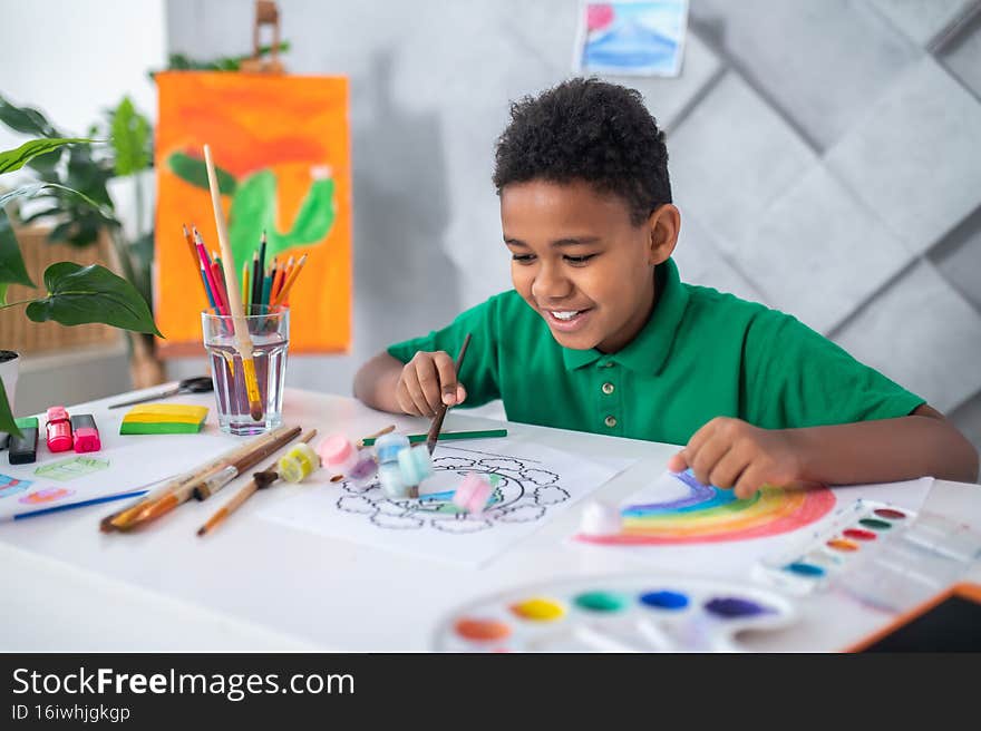 Playful mood. Happy dark-skinned boy of primary school age in green tshirt sitting at table with art supplies pushing tubes of paint standing in picture in bright room. Playful mood. Happy dark-skinned boy of primary school age in green tshirt sitting at table with art supplies pushing tubes of paint standing in picture in bright room