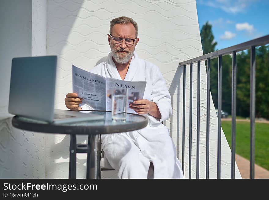 Serious focused man in eyeglasses sitting at the table on the balcony with a newspaper in his hands. Serious focused man in eyeglasses sitting at the table on the balcony with a newspaper in his hands