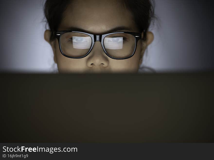 Close-up of concentrated tired Asian woman in glasses looking at computer screen while working indoor at night. Woman surfing Internet. The monitor screen reflected in the glasses. Home Office. Remote work