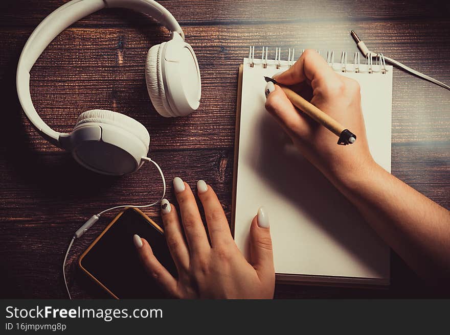 The notebook, the telephone and white headphones are on the table. The woman takes notes in a notebook. The notebook, the telephone and white headphones are on the table. The woman takes notes in a notebook