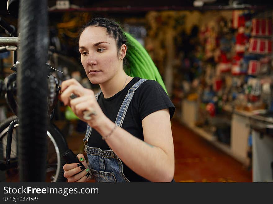 Focused alternative female mechanic with colored dreadlocks using instrument for repairing wheel of bike in workshop. Focused alternative female mechanic with colored dreadlocks using instrument for repairing wheel of bike in workshop
