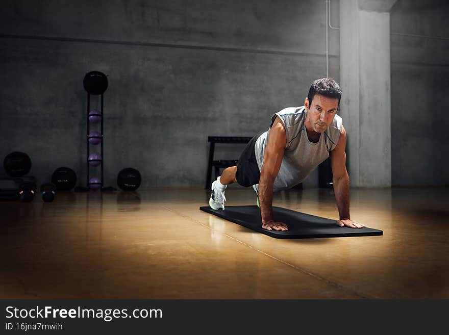 Fitness man in 40s, in grey t-shirt, looks at camera with challenging look, while does push-ups exercises at dark gym floor, indoors. Dramatic dark background. Full shot, Copy-space. Fitness man in 40s, in grey t-shirt, looks at camera with challenging look, while does push-ups exercises at dark gym floor, indoors. Dramatic dark background. Full shot, Copy-space.