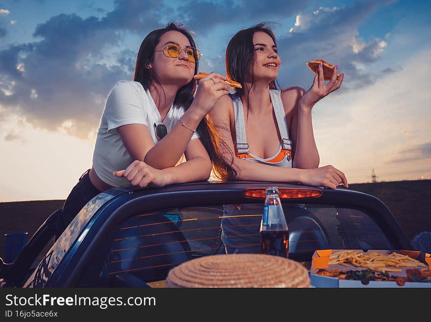 Two women eating pizza, smiling, posing in yellow car cabrio with french fries, hat and soda in glass bottle on trunk