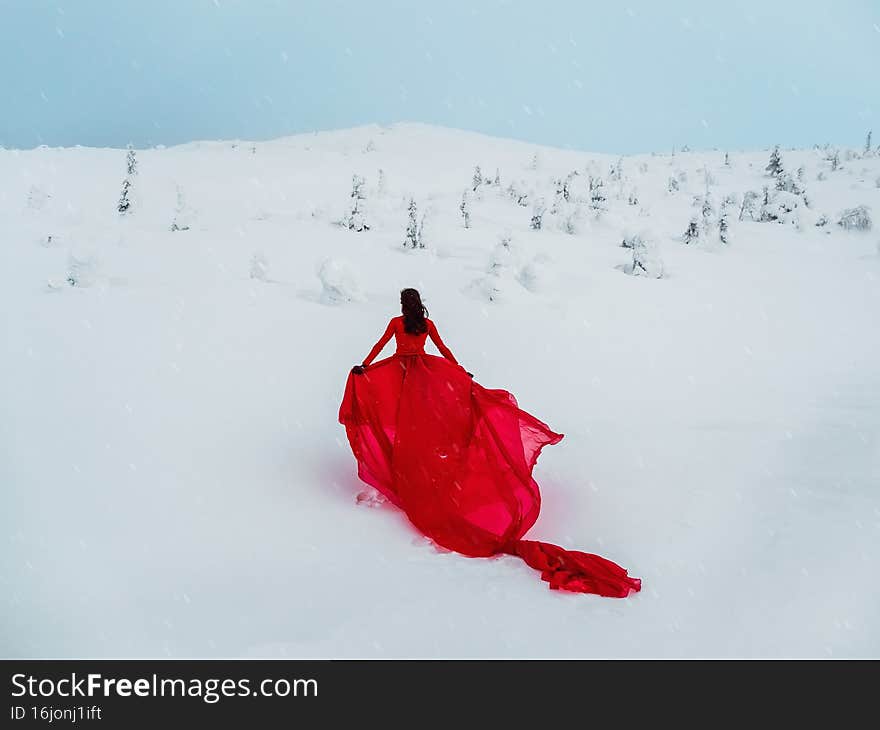 Young beautiful woman in long red dress over winter hill background and snowfall. Fairy tale girl on polar winter landscape. View