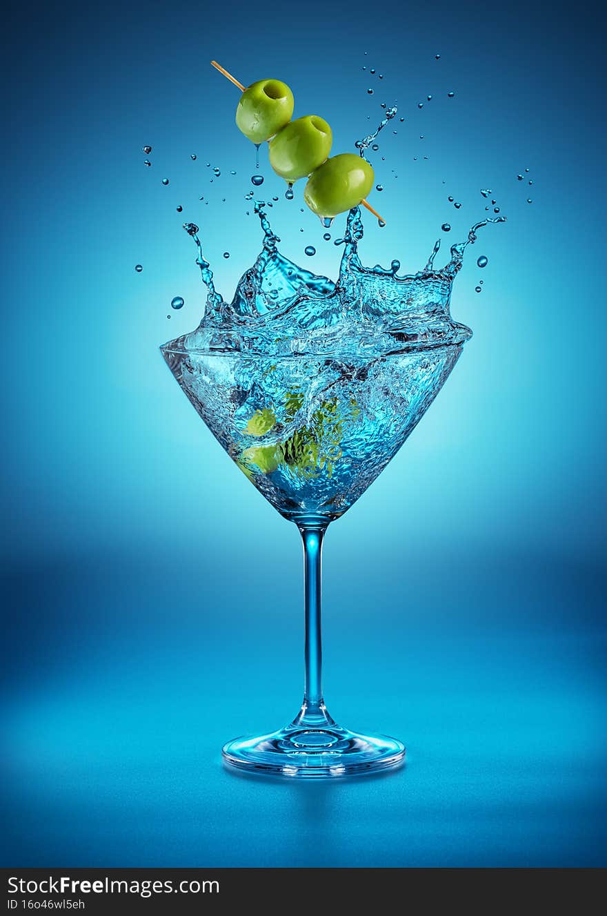 Martini glass with amazing splash as the result of olive berries falling down in it. Blue background