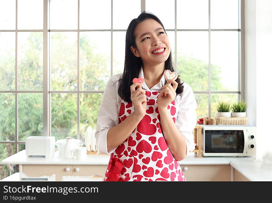 Happy smiling beautiful young Asian woman in cute red heart apron, standing behind kitchen counter, holding valentine homemade cookies, thinking about romantic love memorable anniversary celebration