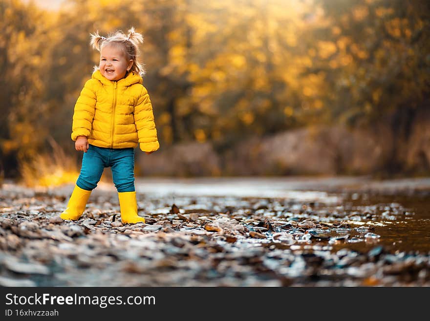 A smiling child girl in yellow rubber boots is playing on the river bank. Copy space. Concept of happy childhood